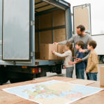 Une femme, un homme et deux enfants se tiennent devant un camion de déménagement moderne, avec des cartons neutres et une carte non annotée de l’Europe posée sur une table.