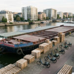 Vue aérienne réaliste d’une péniche de fret amarrée à un quai urbain français, avec des palettes en cours de déchargement et des vélos-cargos électriques prêts à partir, sur fond de ville dense.