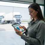 Responsable logistique observant un tableau de bord ESG abstrait sur une tablette dans un entrepôt moderne, avec des camions visibles en arrière-plan et une lumière naturelle.