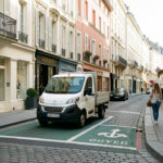 Rue commerçante française calme avec une aire de livraison clairement délimitée, un utilitaire électrique stationné et deux piétons.